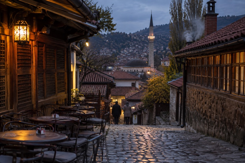 A narrow cobblestone street in an old European town at dusk with empty tables outside a cafe