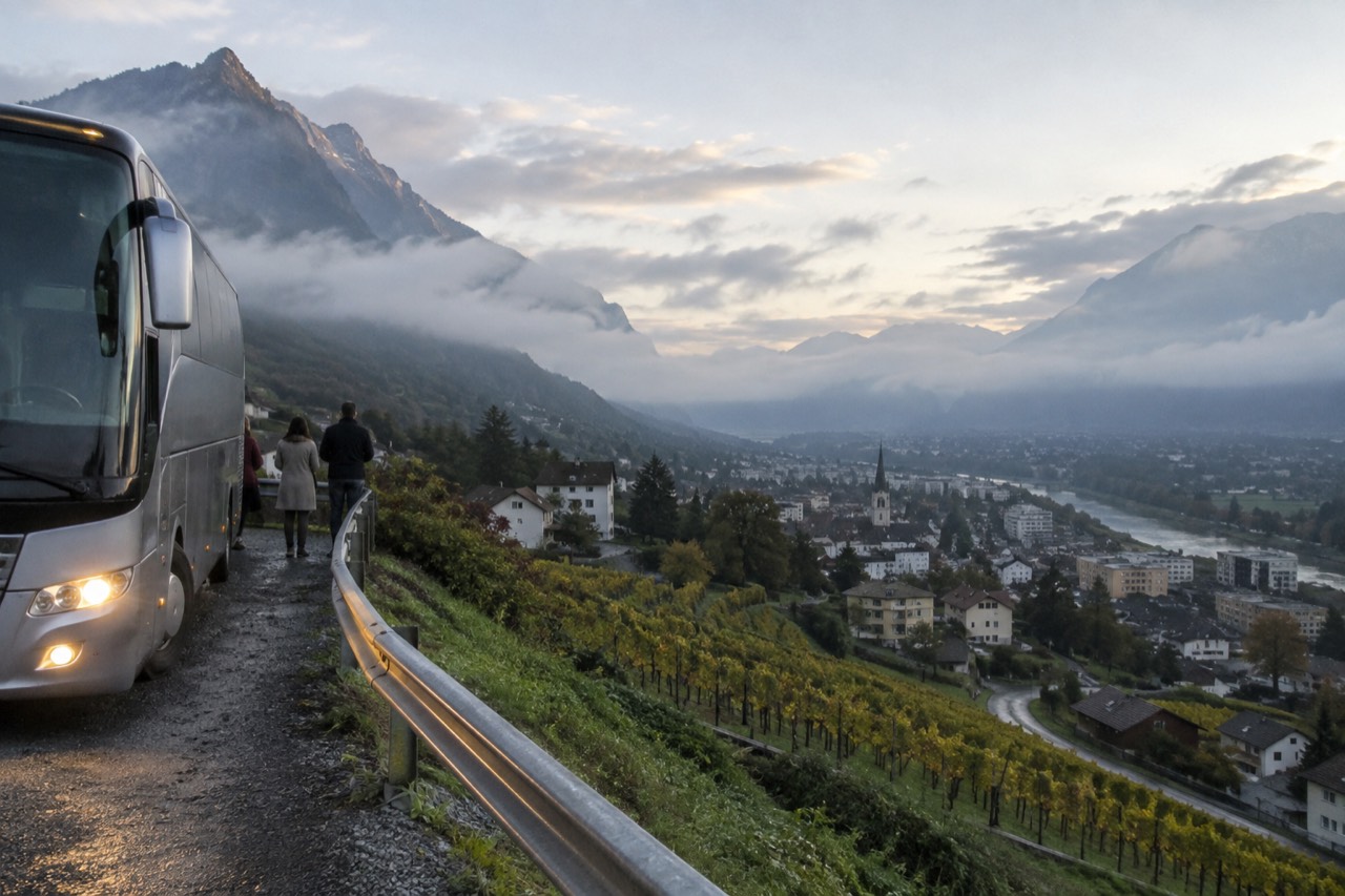A coach parked on a mountain road above a European valley town at golden hour
