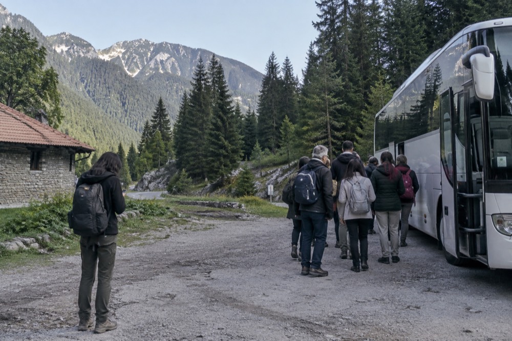A tour bus parked in front of a mountain landscape with passengers boarding, and a separate scene of a solo traveller checking a train timetable on a phone.