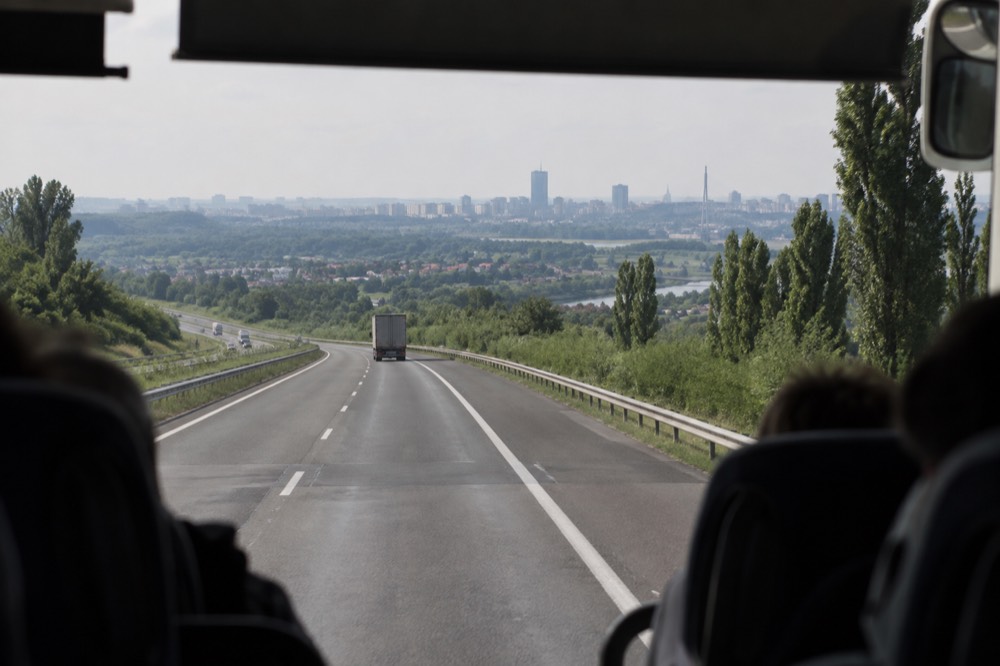 A long distance coach travelling on a highway between green hills toward a distant city skyline