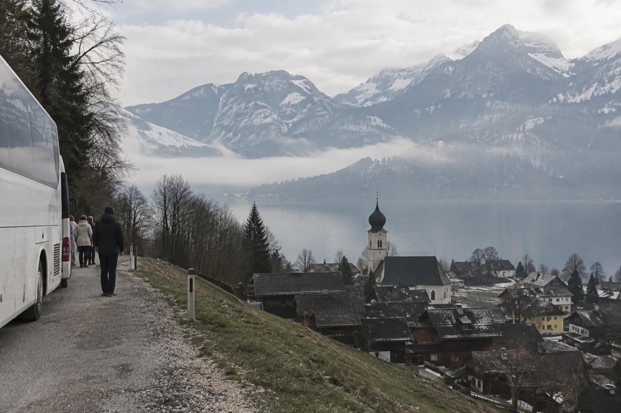 A coach parked on a mountain road above an Alpine valley with village rooftops below
