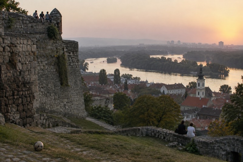 Warm September evening above Belgrade in Serbia, ochre and rust rooftops of the old town where the Sava meets the Danube, hazy light across the water and ancient stone ramparts close by.