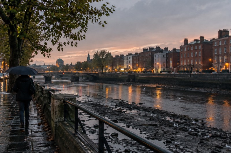 Dusk in Dublin, Ireland, after clearing drizzle, with Georgian red-brick terraces along the Liffey, wet cobbles reflecting amber shoplight, a lone figure under an umbrella and gulls on the low-tide mud.