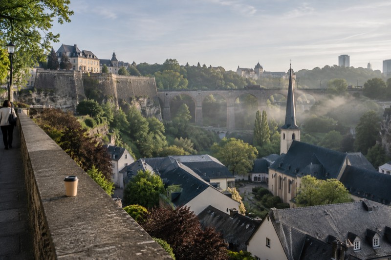 Late spring morning in Luxembourg City, where cream limestone viaduct arches span the wooded Alzette gorge, mist lingering below and a church spire rising over tiled rooftops.