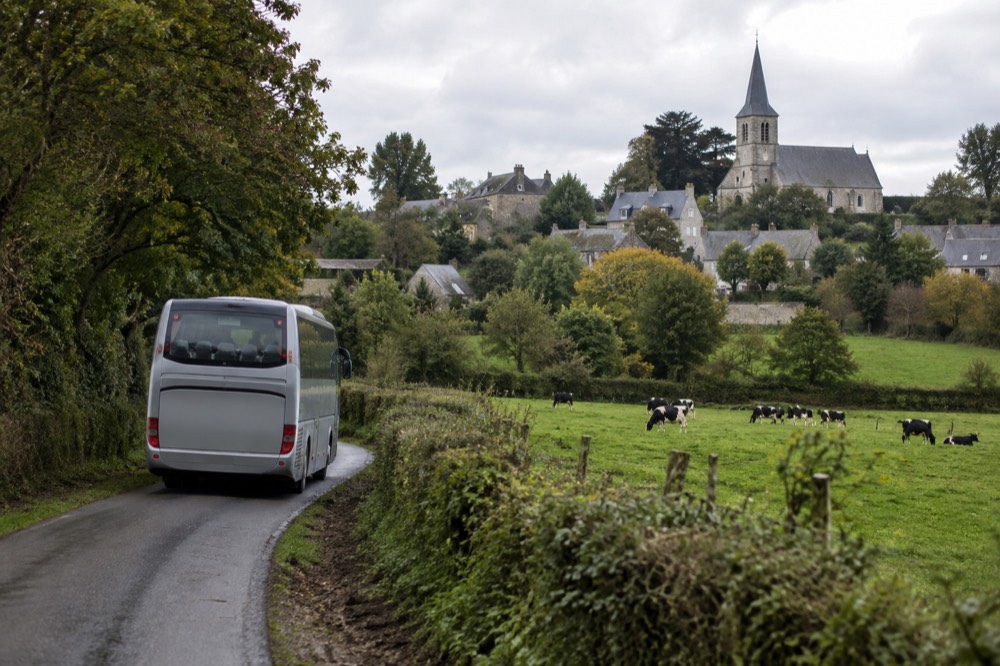 A touring coach on a road winding through green European countryside with a village church in the distance