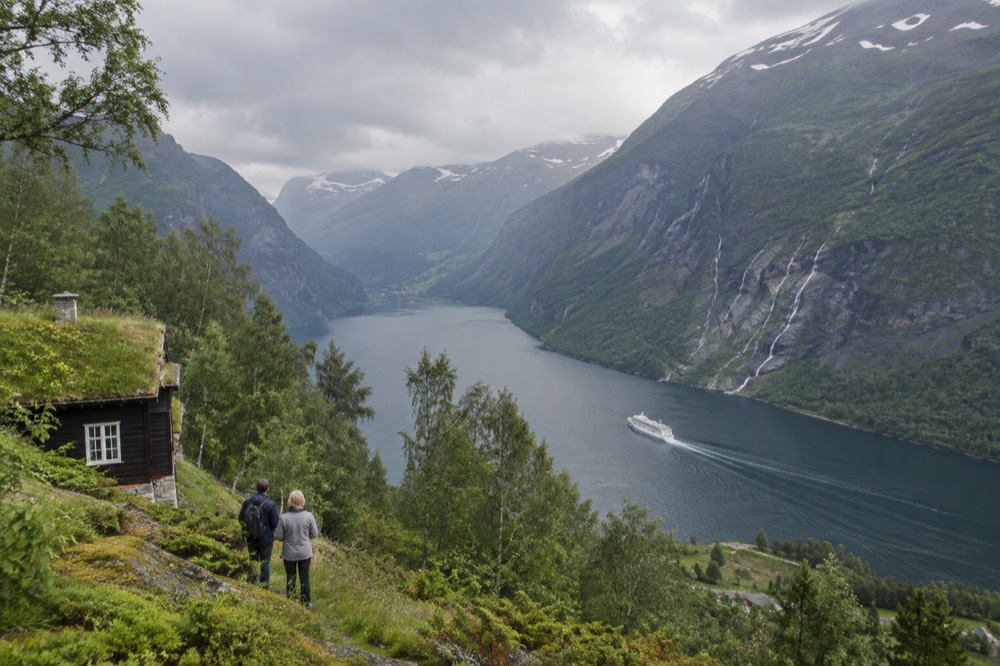 Steep green cliffs rising above a deep blue Norwegian fjord with a small cruise ship in the distance
