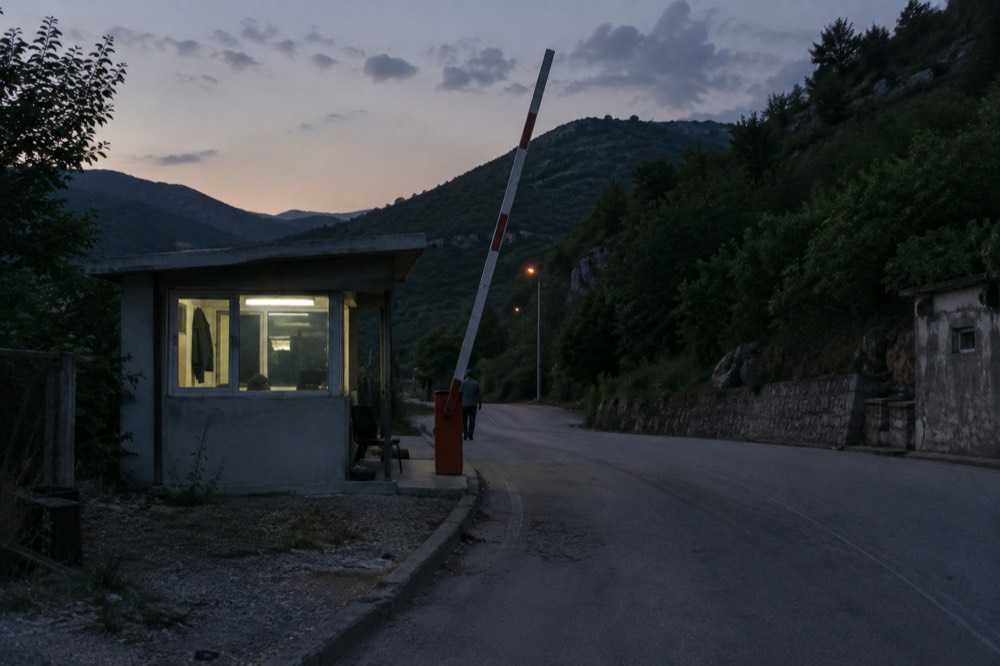 A small border crossing station on a road between green hills at dusk
