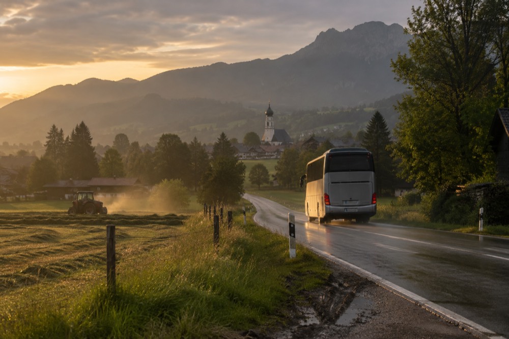 A touring coach on a road between rolling green hills with a distant village church spire