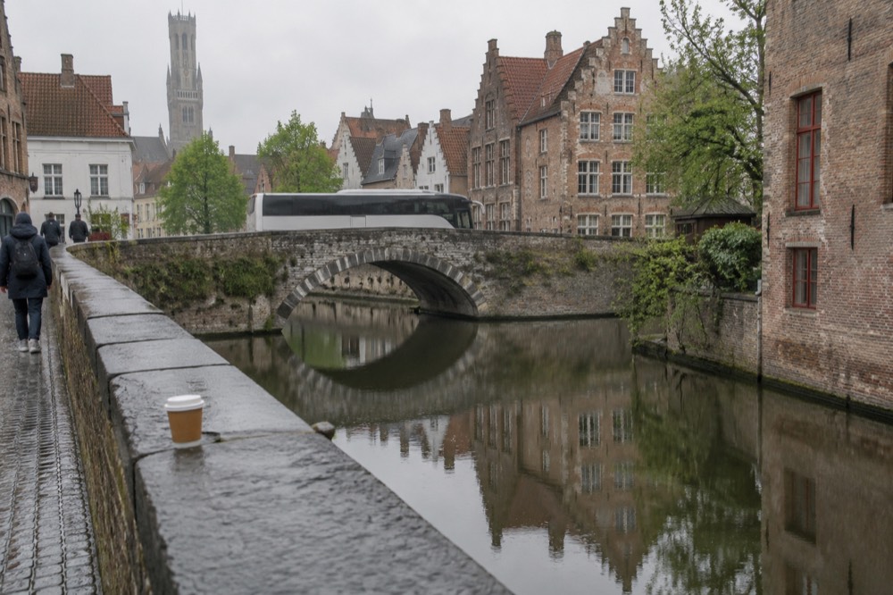 A touring coach on a road beside a river with a historic stone bridge and old town buildings