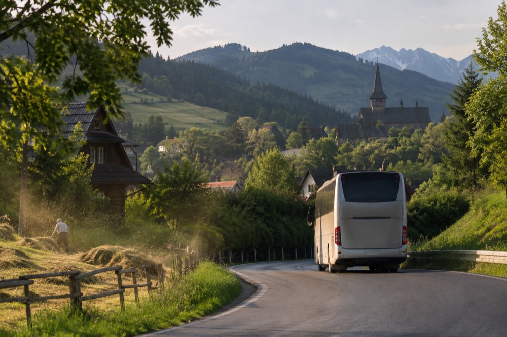 A coach travelling a curving road through green hills with a distant village church spire