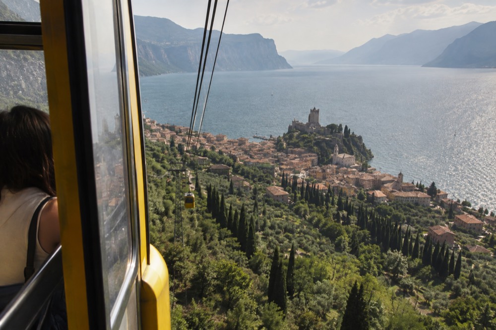 A cable car cabin rising over a forested slope with a lake and town visible below