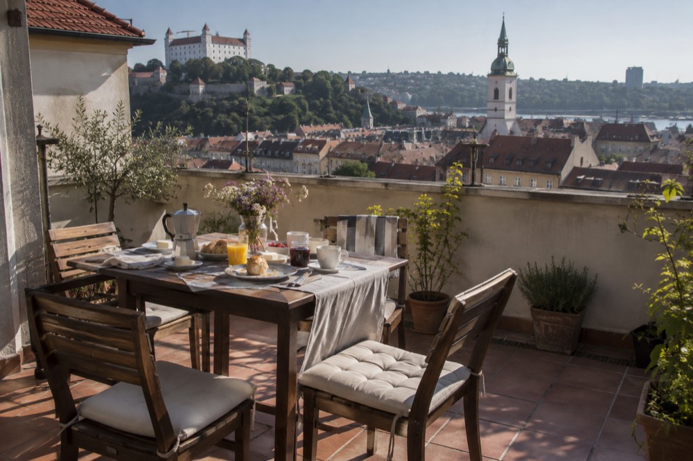 A family-sized dining table set for breakfast on a terrace overlooking a European old town