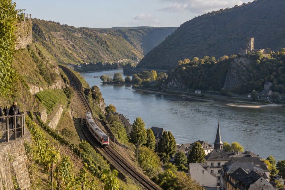 A train winding along a hillside line above a river with a historic town on the opposite bank