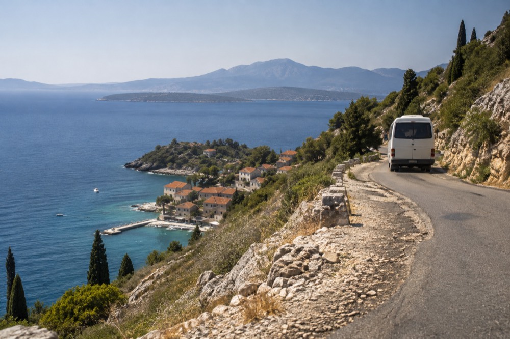 A small minibus on a coastal road beside turquoise Adriatic water
