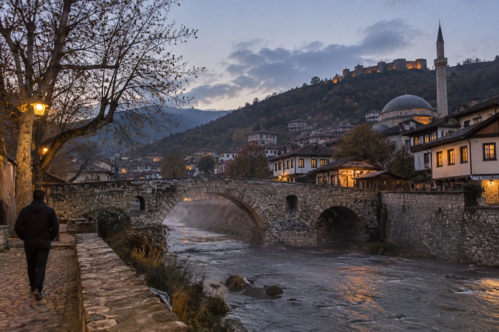 A stone bridge over a river in a Balkan city at dusk with lit windows along the bank