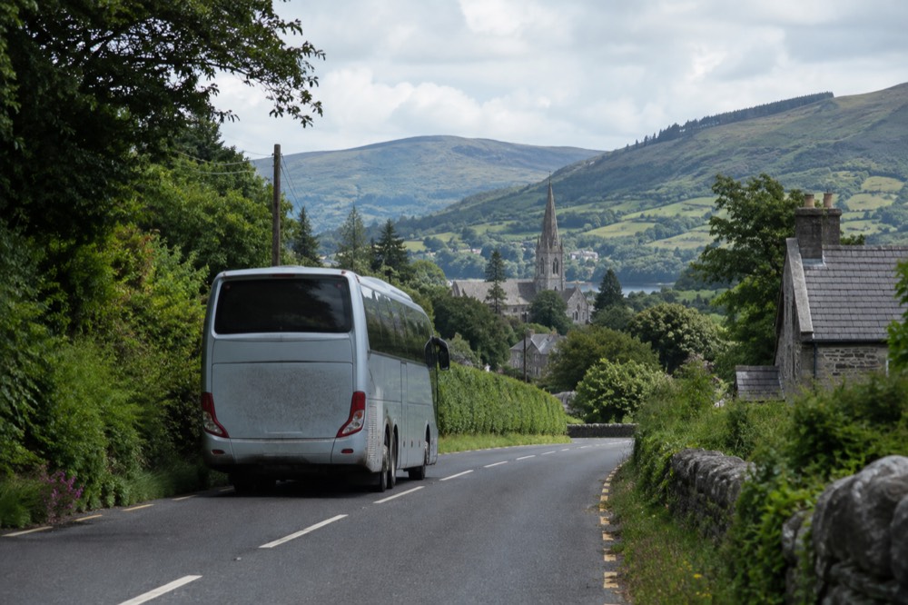 A touring coach driving on a road between green hills with a distant village church spire